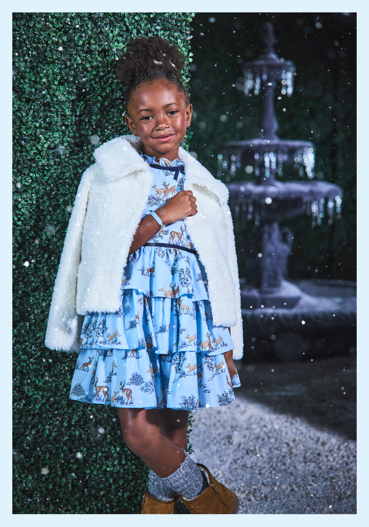 Young girl wearing a blue deer-print tiered dress and white faux fur jacket, posing in a snowy garden setting