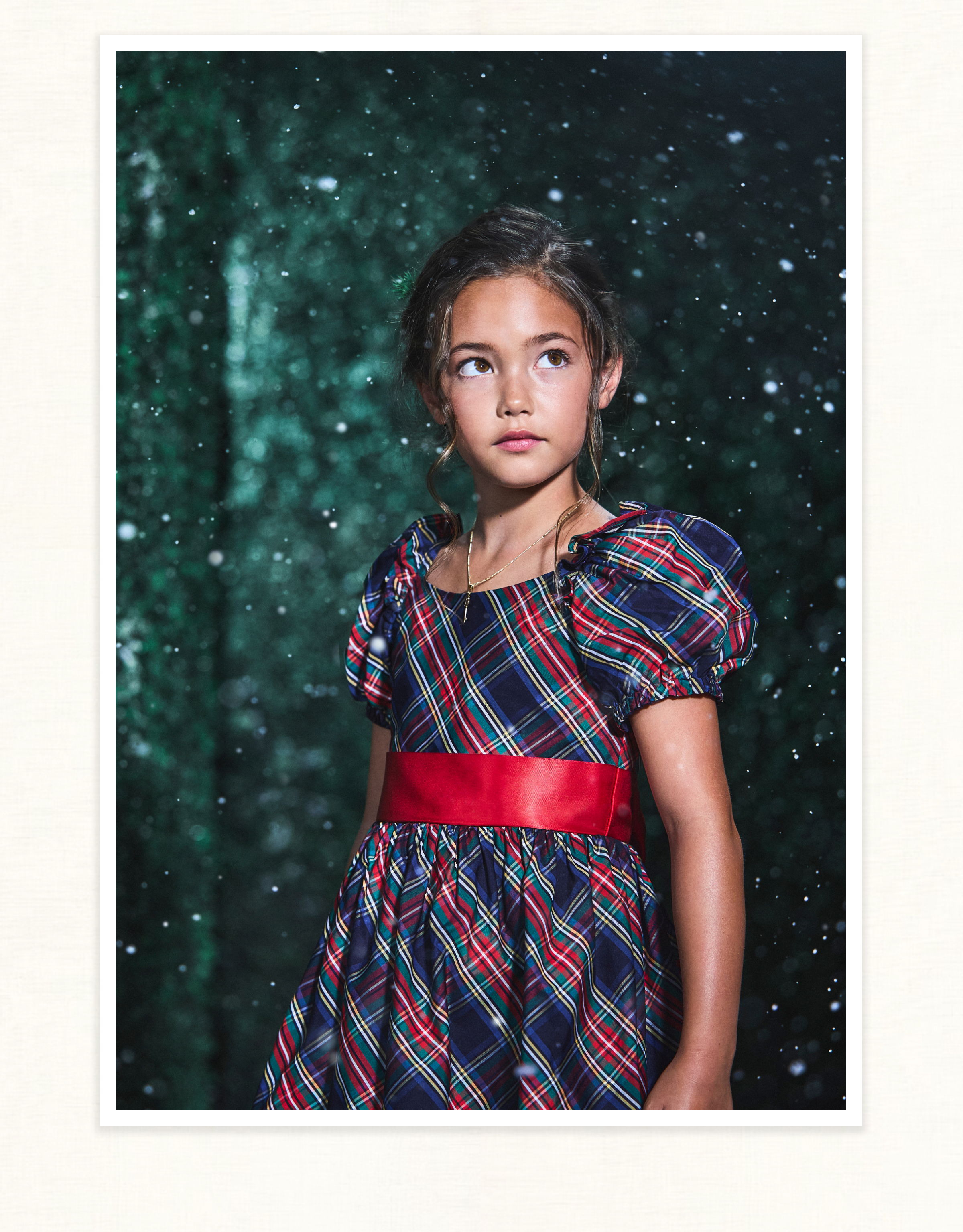 Young girl in tartan plaid dress with red sash, standing in a snowy forest backdrop