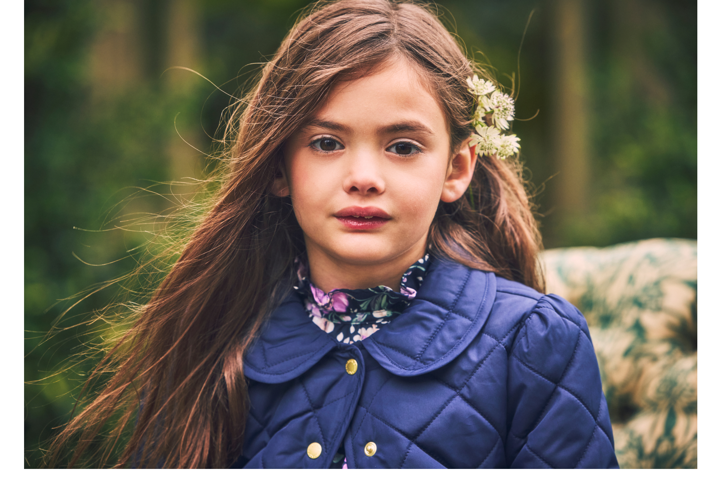 Young girl with long brown hair and white flowers, wearing a navy quilted jacket over a floral blouse
