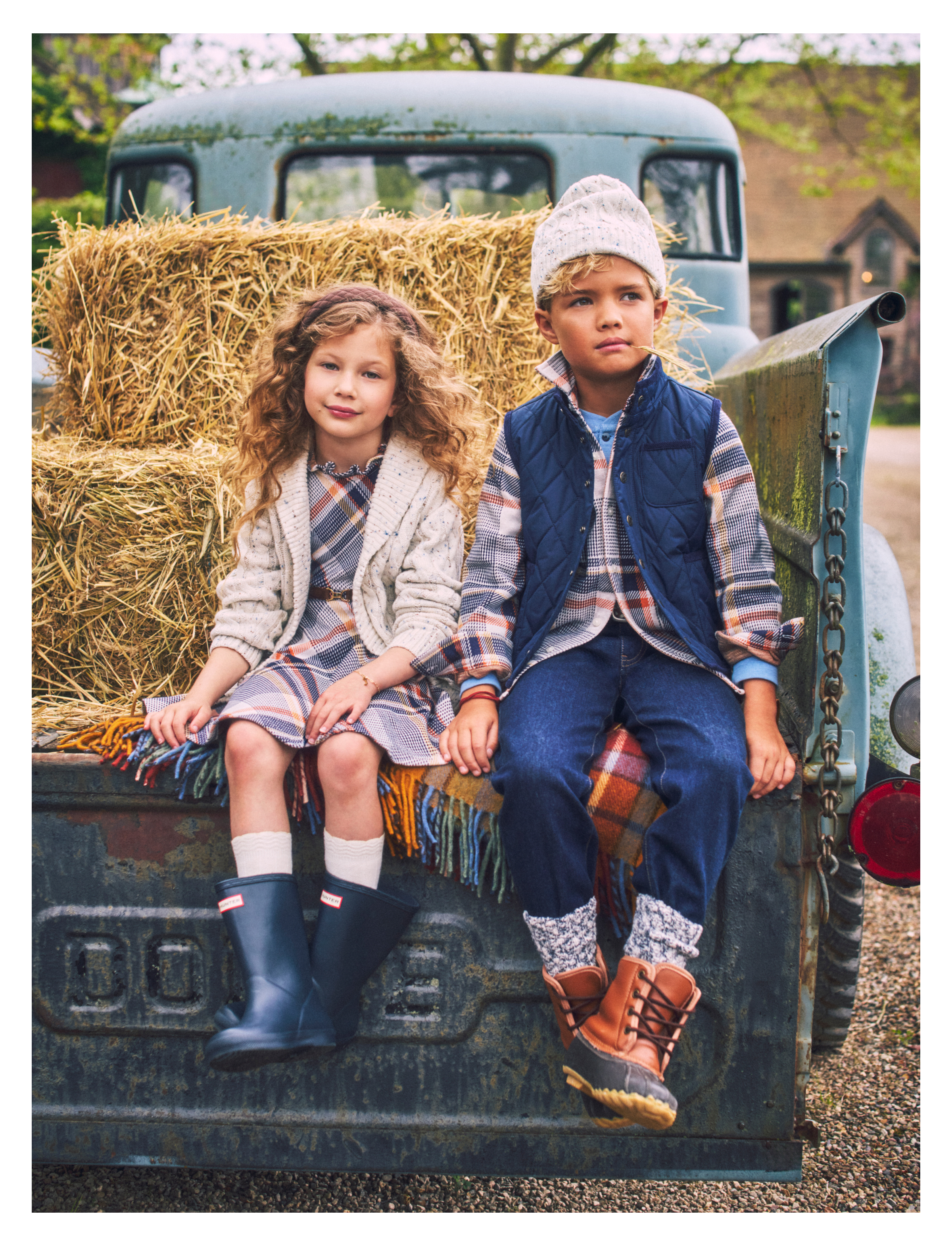 GIRL AND BOY DRESSED IN FALL CLOTHING SITTING ON BACK OF OLD TRUCK WITH BUNDLES OF HAY