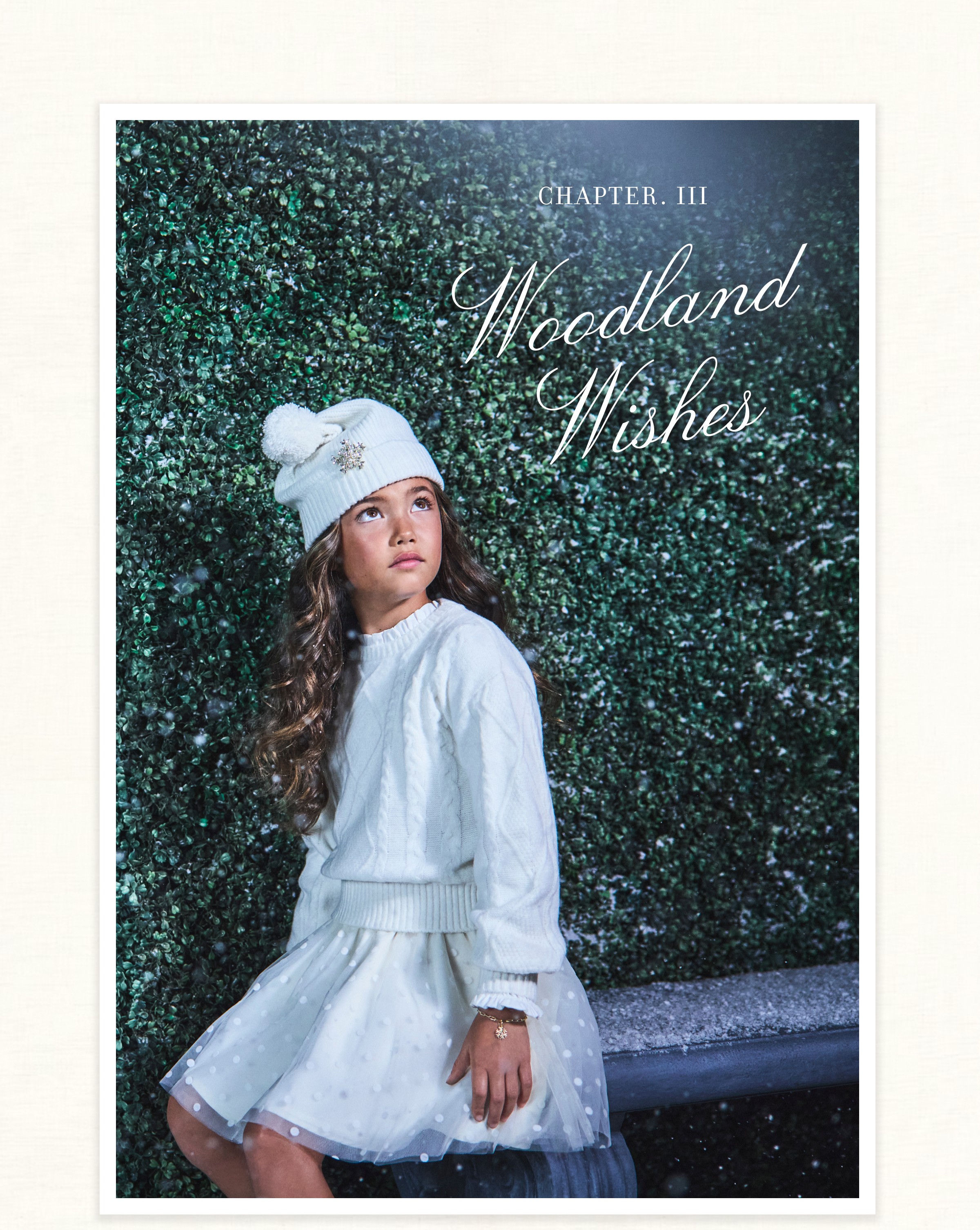 Girl in white cable-knit sweater, tulle skirt, and pom-pom beanie against a green hedge with falling snow - Chapter. III - Woodland Wishes