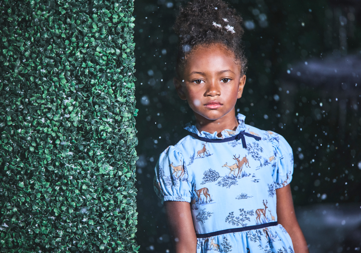 Young girl in blue deer-print dress standing beside a green hedge wall in falling snow