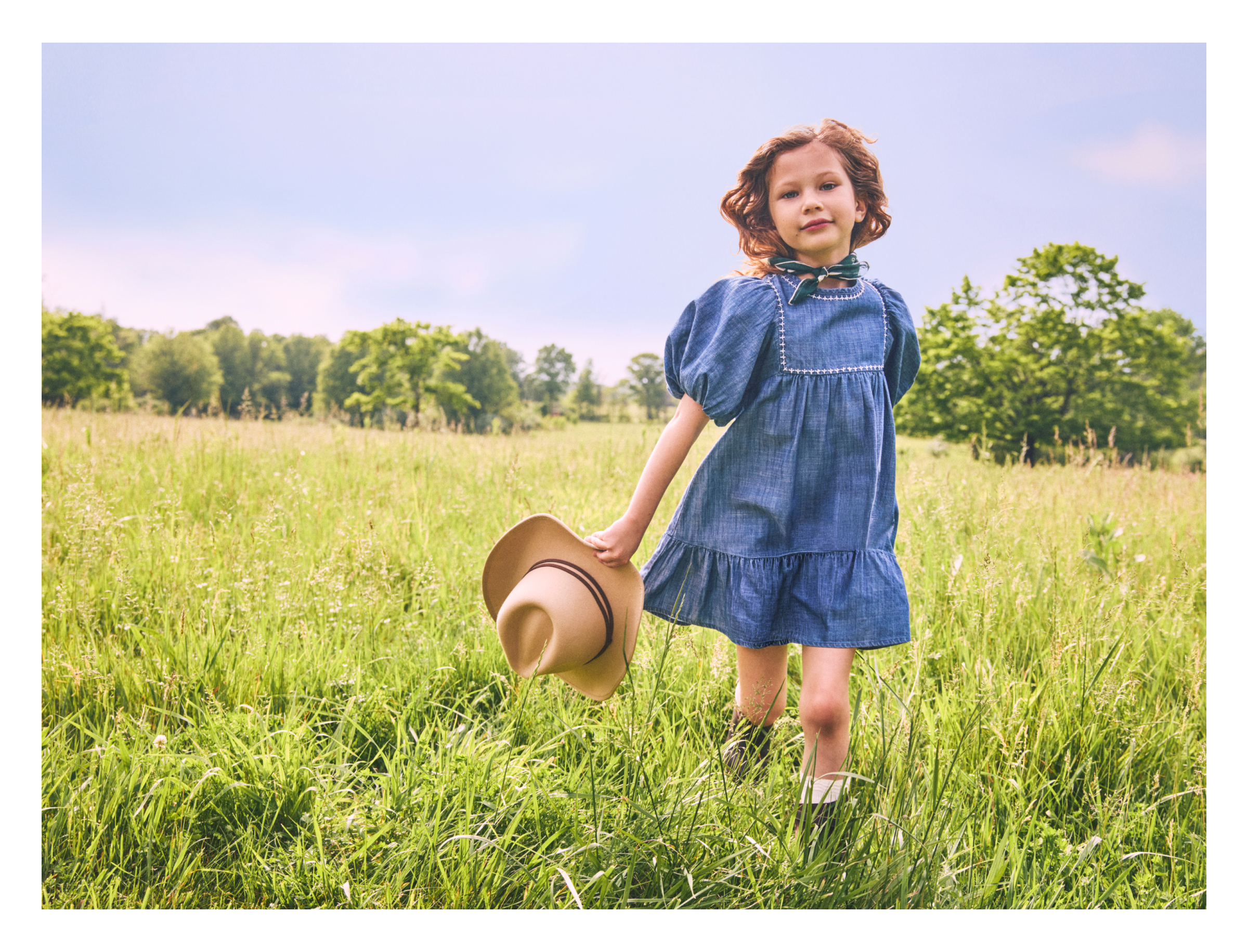 GIRL IN DENIM DRESS HOLDING WESTERN HAT WALKING IN GRASSY FIELD