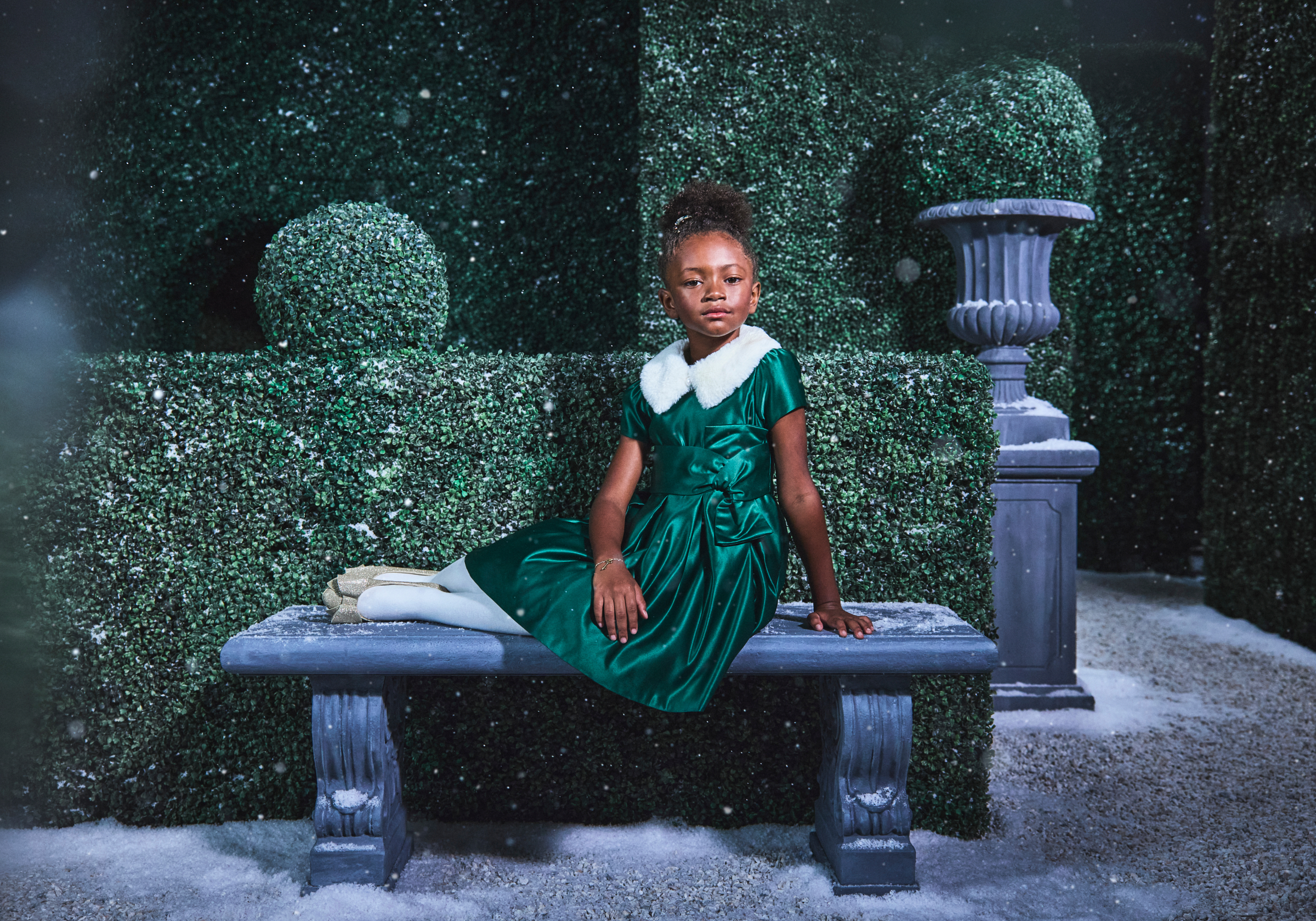 Young girl in emerald satin dress with white fur collar seated on stone bench in snowy garden