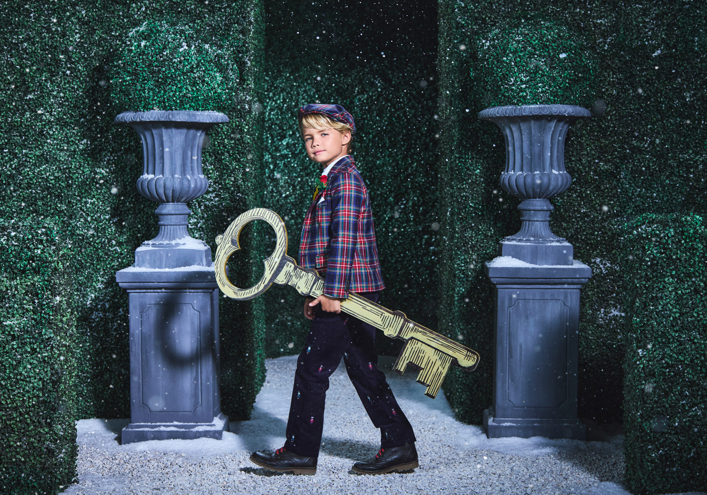 Boy in plaid blazer and cap carrying oversized decorative key through snowy hedge maze