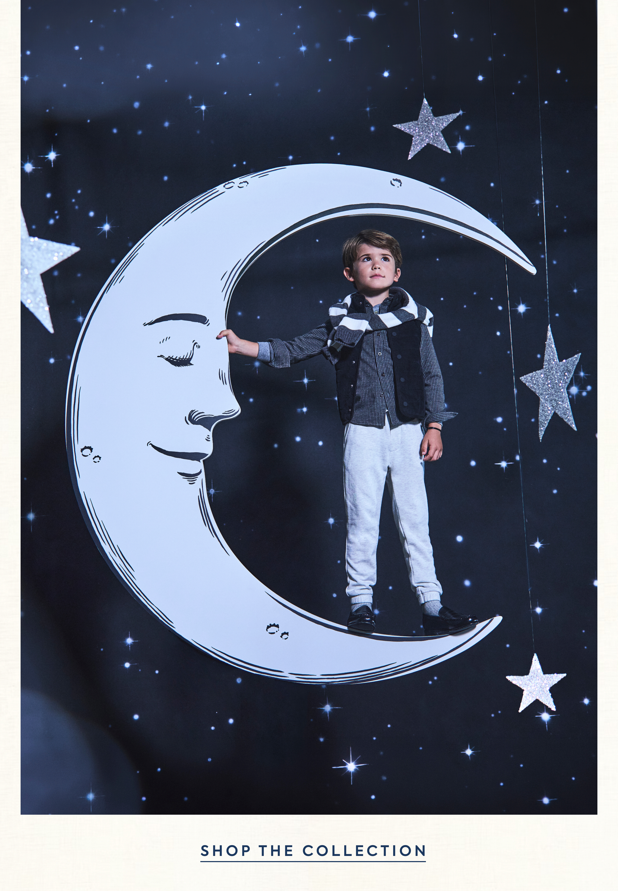 Boy standing on a crescent moon prop in a starry night scene wearing a vest, striped scarf, and grey pants - Shop the Collection