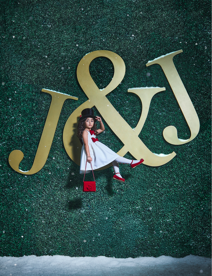 GIRL IN WHITE DRESS WITH RED BOWS AND A BLACK TOP HAT SITTING ON A JANIE AND JACK SIGN