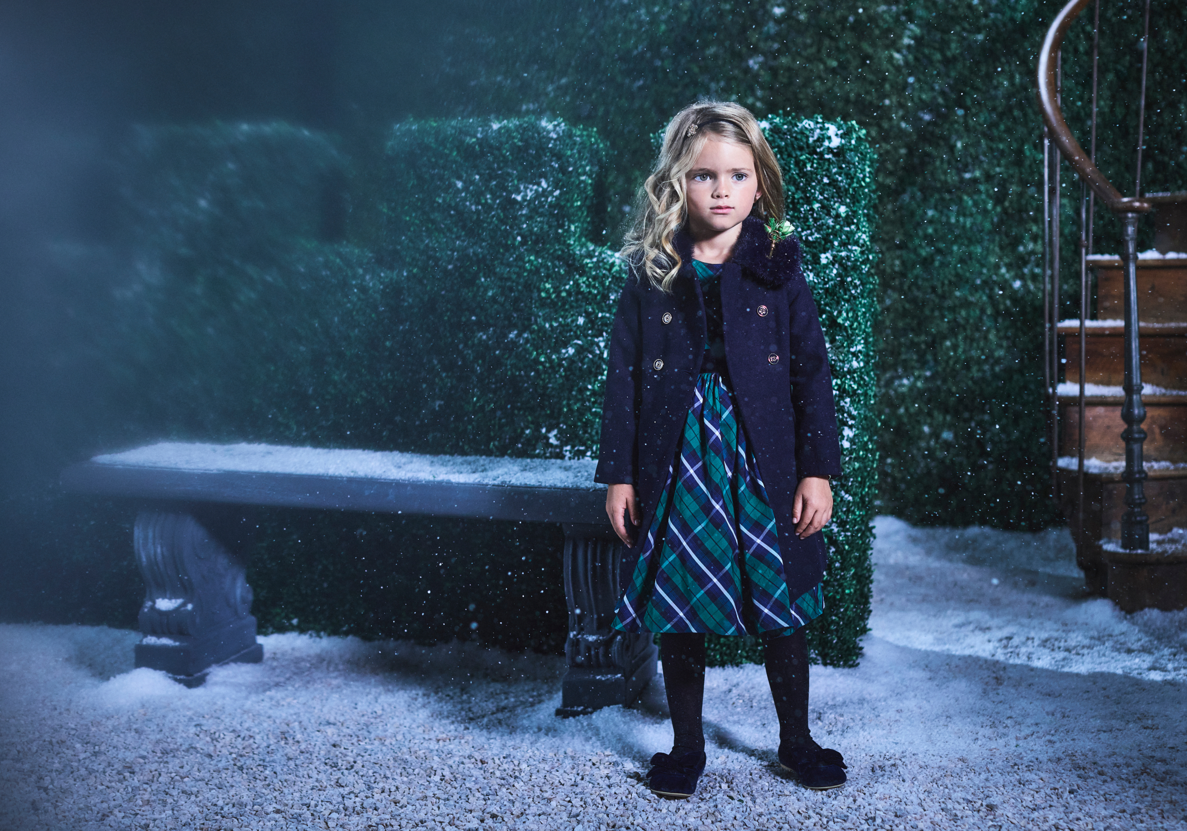 Young girl in navy coat and green plaid dress standing in a snowy garden at night