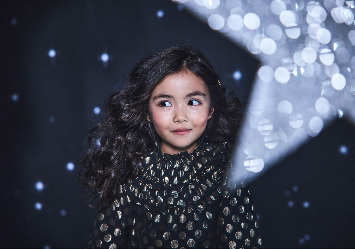 Young girl in black and gold polka dot dress under sparkling lights - All the Trimmings