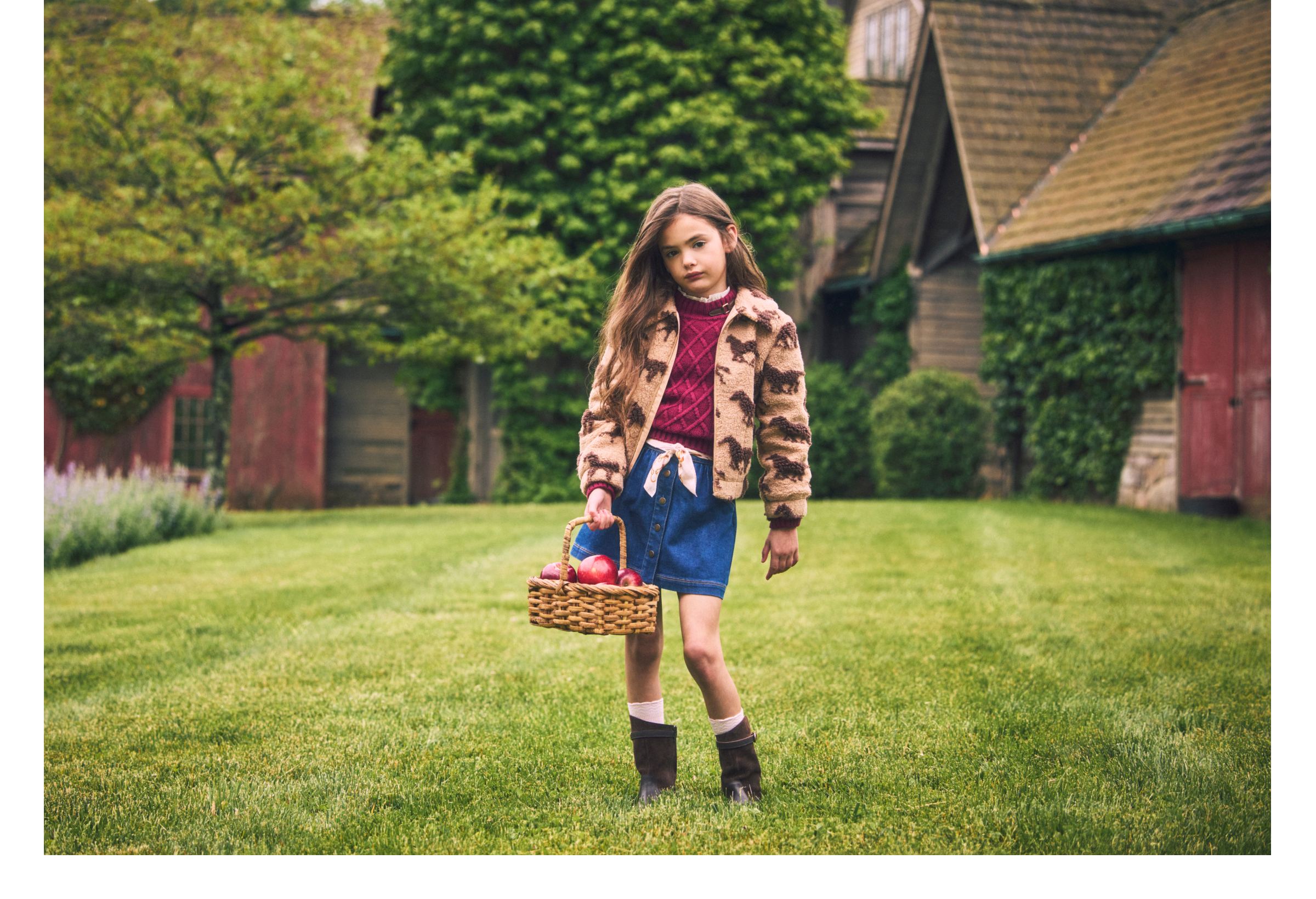 Girl in horse-print jacket and denim skirt carrying a basket of apples on a farm lawn