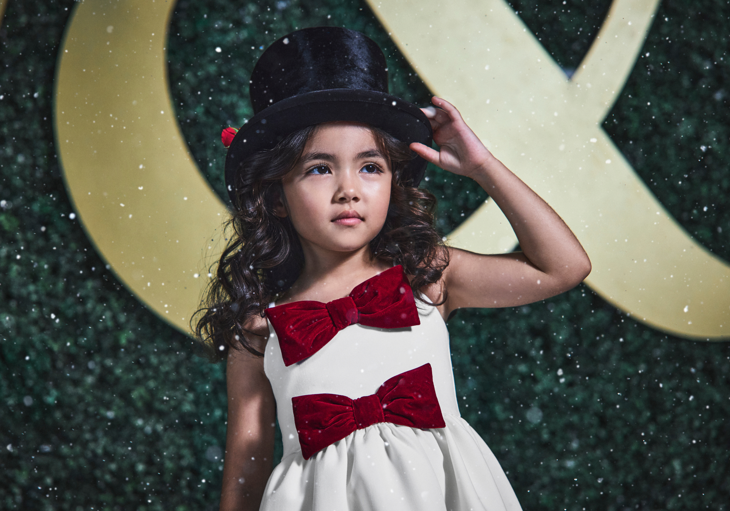 Young girl in white dress with red velvet bows and black top hat posed against green hedge backdrop with falling snow