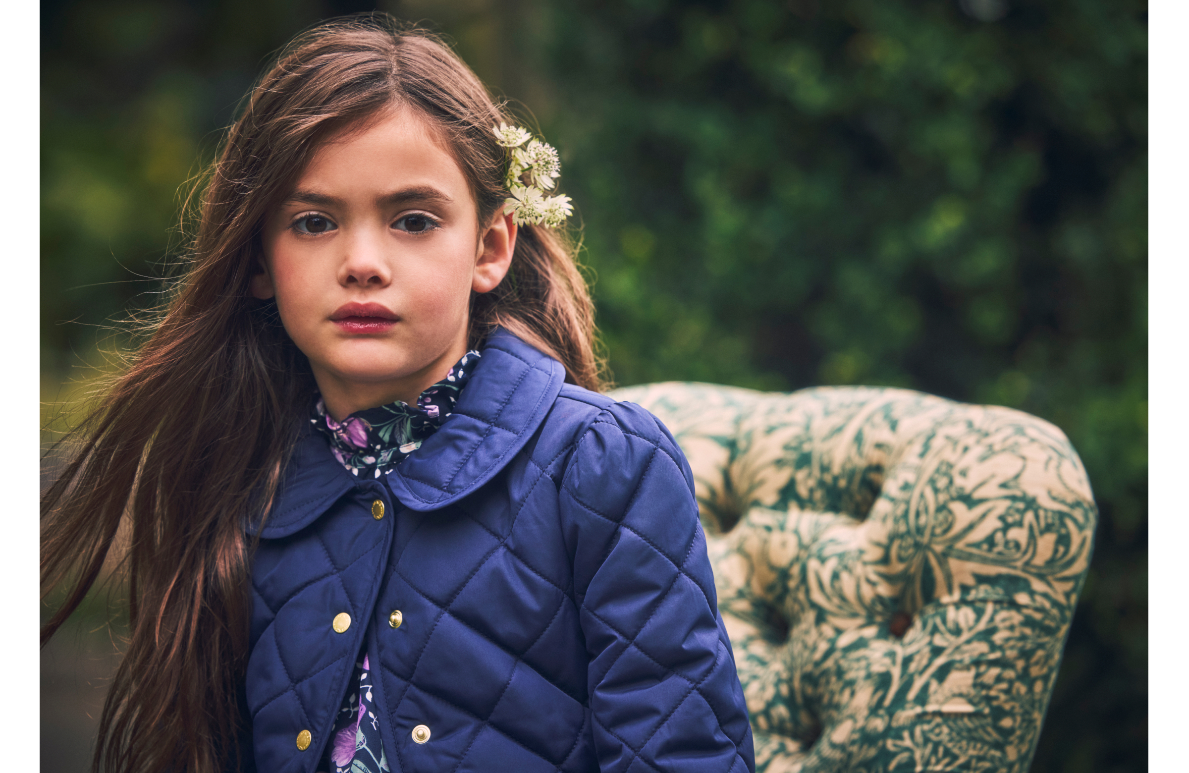 Young girl in navy quilted jacket with floral hair accessory seated near ornate green and cream chair