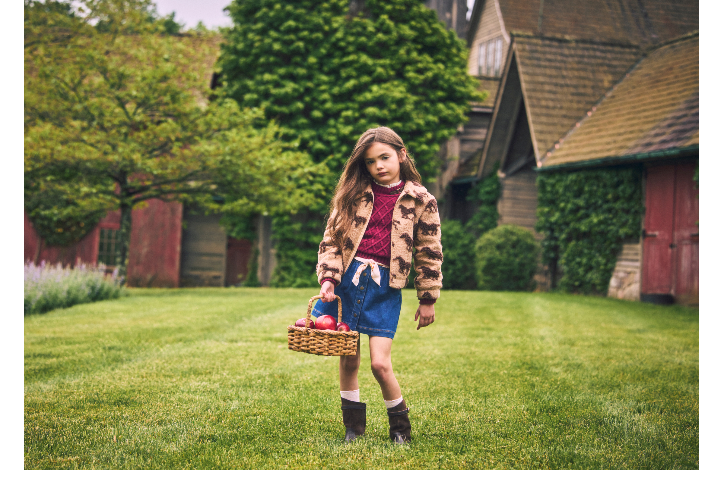 Young girl in horse-print jacket and denim skirt carrying a basket of apples on a farm lawn