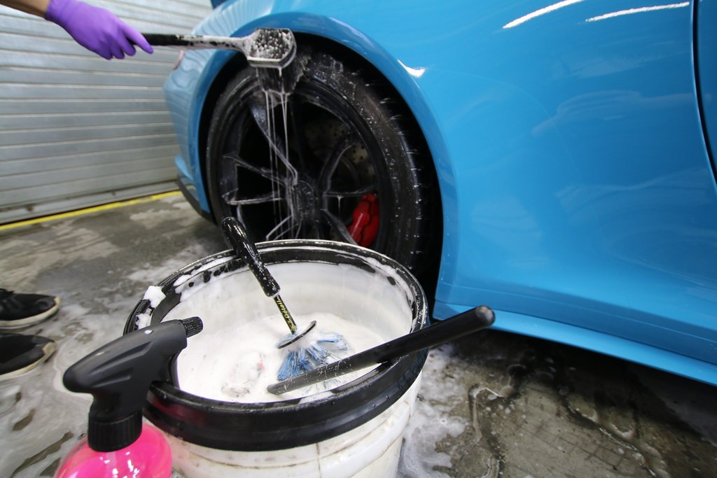 Wash bucket and wheel brush beside the front wheel of a blue Porsche 911 in the Detailer's Domain shop