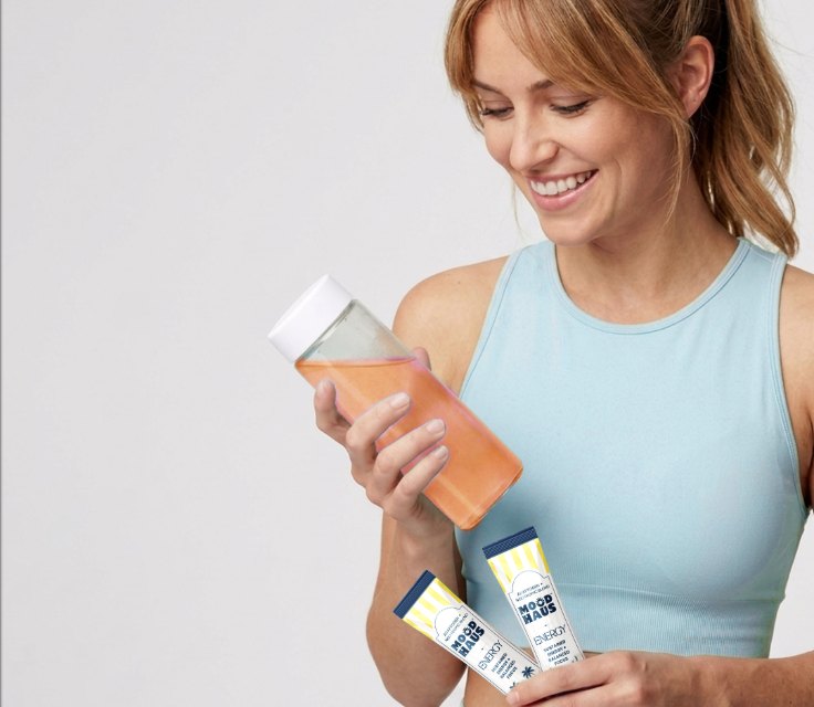 A smiling woman in activewear holds a bottle of ENERGY and two stick packs
