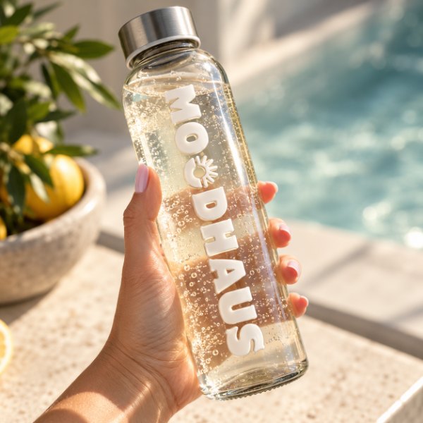 A hand holds the MOODHAUS glass water bottle by the pool