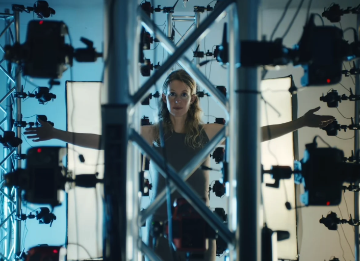 A woman with long blonde hair stands with arms outstretched surrounded by cameras and sensors.