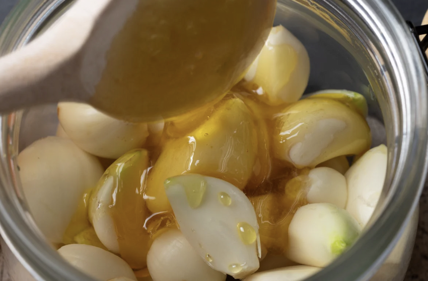 A spoon drips honey onto many garlic cloves in a jar