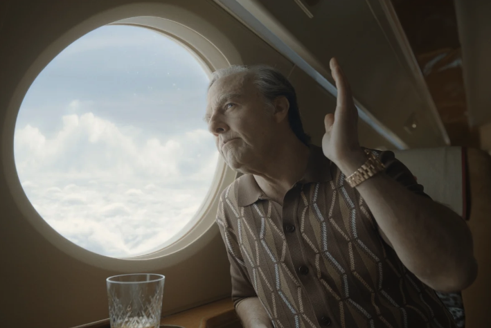 Tom Cruise in character as Digger Rockwell looking out a plane window with one hand raised.