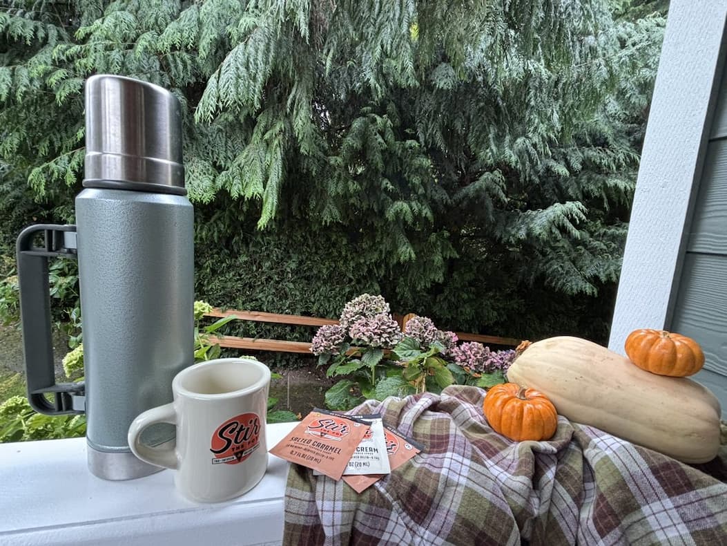 A green thermos and a Stir The Pot mug on a porch rail with Salted Caramel and Vanilla Cream sachets, a plaid blanket, squash and small pumpkins