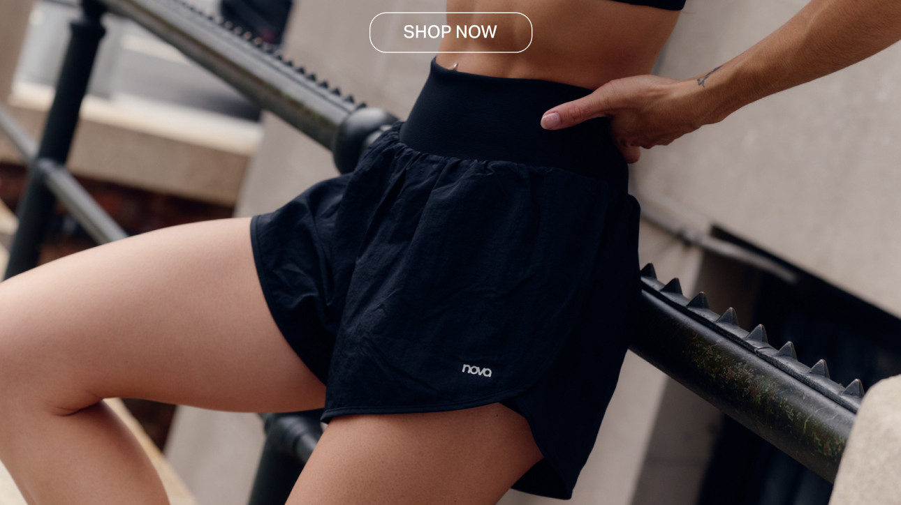 Close-up of black Nova high-waisted athletic shorts on a person posing by a railing, featuring a Shop Now prompt to buy women's activewear.