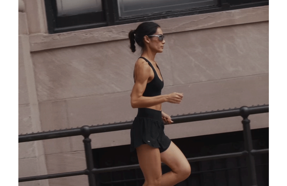 Woman running in an urban setting wearing a black sports bra and matching shorts, showcasing new women's athletic running apparel.